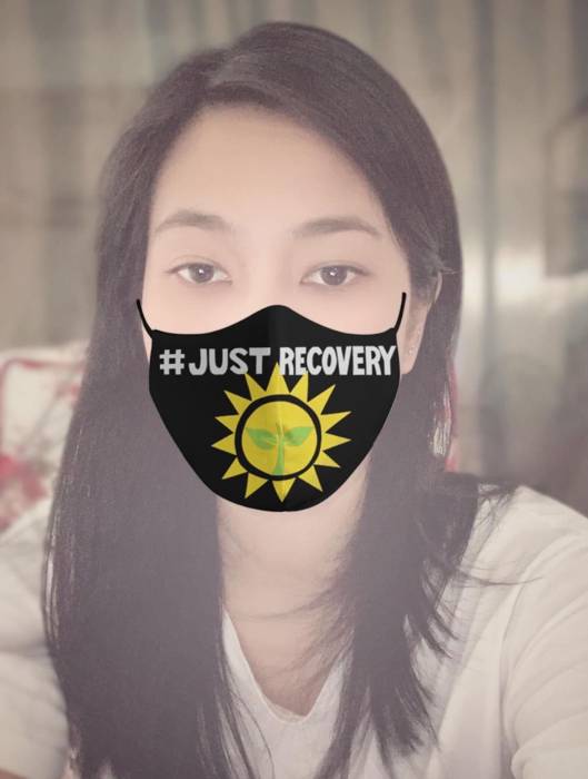 A photo of a woman with a black facemask with white letters reading Just Recovery and a symbol of the sun